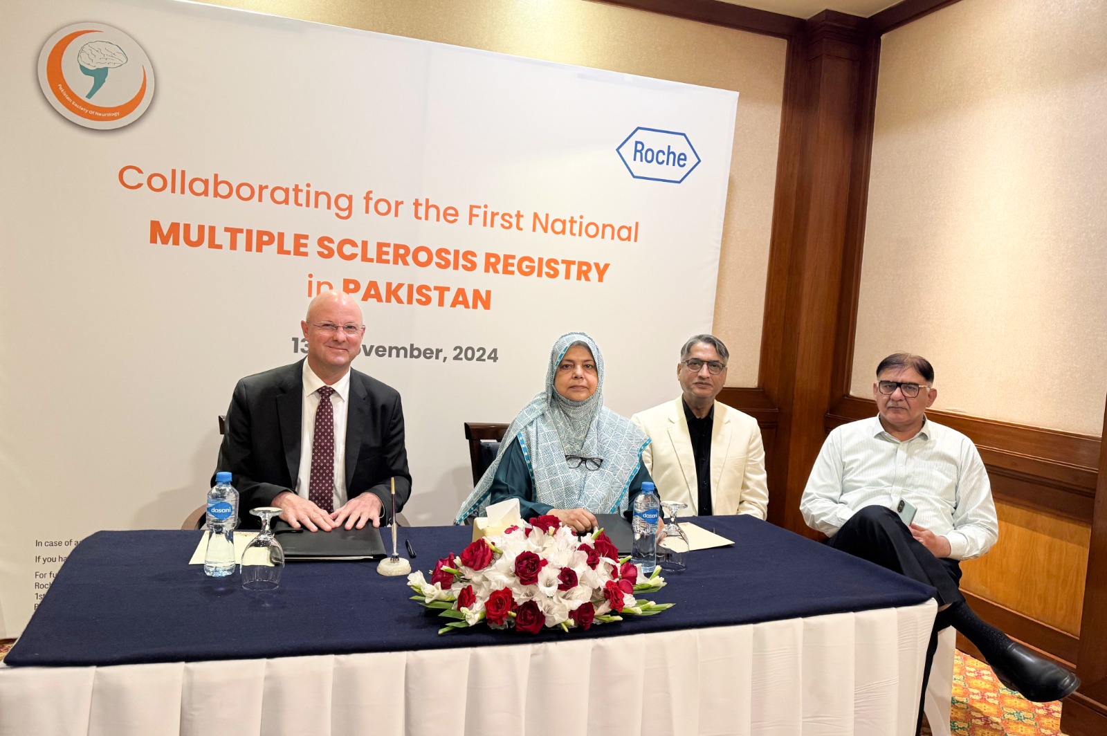 A group of people at a signing event for the National Multiple Sclerosis Registry in Pakistan. Two individuals are seated, signing documents, while others stand behind them. The mood is formal, with a logo and text in the background.