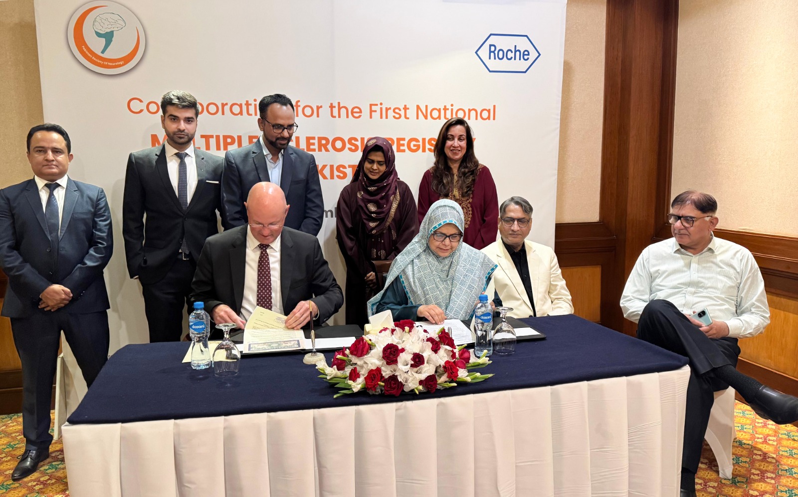 A group of people at a signing event for the National Multiple Sclerosis Registry in Pakistan. Two individuals are seated, signing documents, while others stand behind them. The mood is formal, with a logo and text in the background.