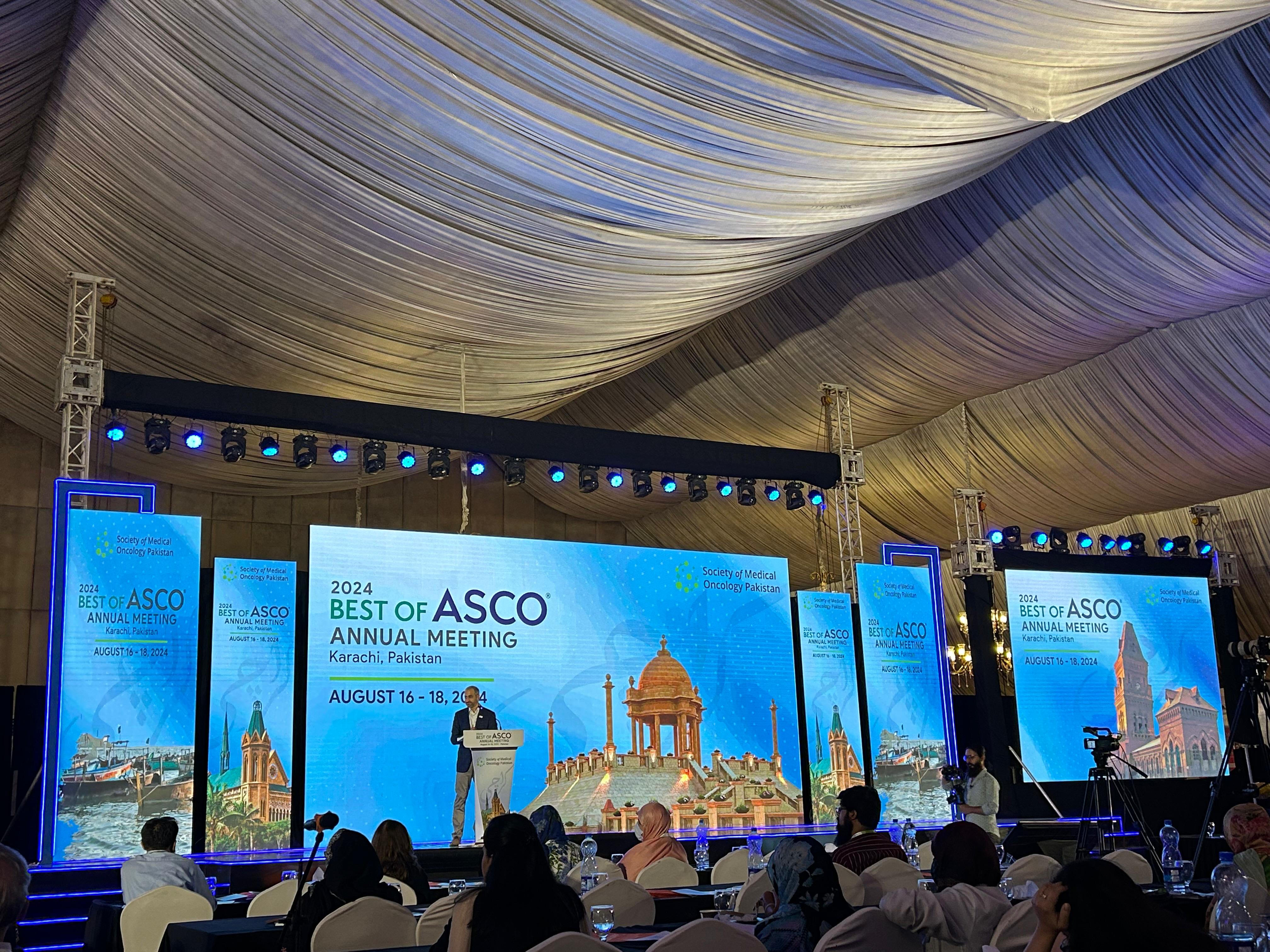 A speaker presents at the "2024 Best of ASCO Annual Meeting" in Karachi, Pakistan. The venue has a large screen with city landmarks and an attentive audience.