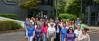 A diverse group of smiling people, wearing colorful shirts, walk together outside a modern building with a Roche logo. The mood is cheerful and collaborative.