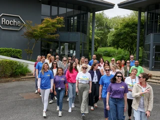 A diverse group of smiling people, wearing colorful shirts, walk together outside a modern building with a Roche logo. The mood is cheerful and collaborative.