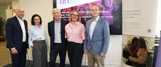 Five people stand smiling in an office setting. Behind them, a screen displays a Healthcare Businesswomen’s Association presentation. A banner features healthcare innovation text.