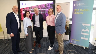 Five people stand smiling in an office setting. Behind them, a screen displays a Healthcare Businesswomen’s Association presentation. A banner features healthcare innovation text.