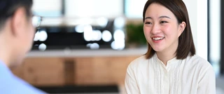 Smiling woman in a white blouse sits facing a blurred person in a blue shirt, in a bright office. The scene conveys a friendly, positive atmosphere.
