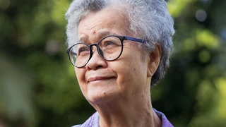 Imagem de uma mulher asiática na terceira idade. Ela possui cabelos grisalhos e usa um óculos com armação preta arredondada. Ela está em um local aberto e arborizado e olha para o horizonte.