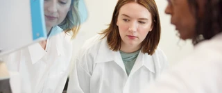 Tre ricercatrici in camice bianco che collaborano in un laboratorio, esaminando e discutendo attentamente dei dati.