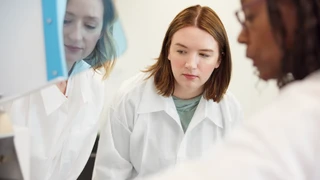 Tre ricercatrici in camice bianco che collaborano in un laboratorio, esaminando e discutendo attentamente dei dati.