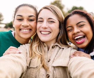 Three women take a joyful selfie outdoors, smiling widely. They stand close together, conveying warmth and friendship in a bright, natural setting.