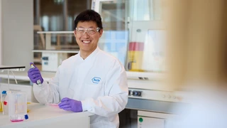 Smiling scientist in a lab coat and safety glasses holds a pipette in a modern laboratory. The setting conveys focus and innovation.