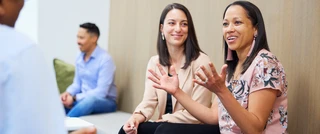 A diverse group of four people sits on a light bench against a wooden wall, engaged in a lively conversation, expressing warmth and connection.
