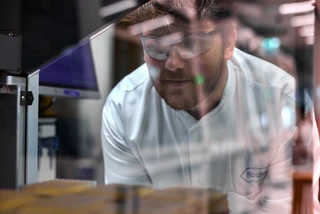 A male scientist wearing safety glasses and a white Roche lab coat observes samples inside a lab instrument, reflecting his focus and the lab environment around him
