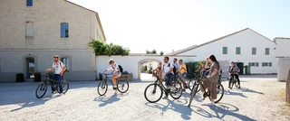 A group of people riding bicycles on a sunny day, in front of rustic stone buildings. The scene conveys a sense of leisure and camaraderie.