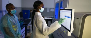 Two lab technicians in protective gear and face masks work in a lab. One operates a touchscreen on a large machine. The setting appears focused and clinical.