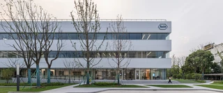 Modern office building with a sleek design, large windows, and minimalistic landscaping. A few young trees are in front, and the sky is overcast.
