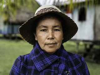 A woman in a straw hat and blue plaid jacket stands outdoors, with a calm expression. Soft background of greenery and a rustic building.