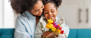 A woman and her daughter sit on a couch, both smiling while holding a bouquet of colorful flowers.