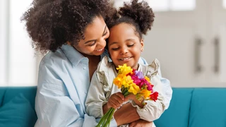 A woman and her daughter sit on a couch, both smiling while holding a bouquet of colorful flowers.