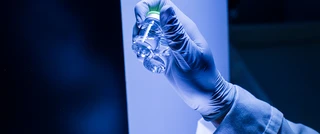 A person in a lab coat holds a glass bottle, standing in a laboratory setting with equipment in the background.