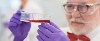 Elderly scientist with glasses and mustache examines red liquid in a petri dish. He wears purple gloves and a red bow tie, conveying focus and precision.