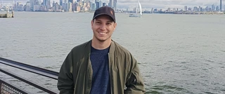 Jorge Rodriguez stands on a bridge, wearing a hat and jacket, with a vibrant city skyline in the background.