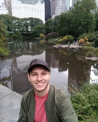 Jorge Rodriguez smiles warmly in front of a serene pond, surrounded by lush greenery in a tranquil park setting.