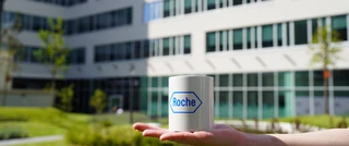 A person holding a cup featuring the logo of Roche, in front of the Budapest RSS office.