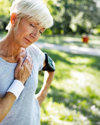 An elderly woman with short white hair, wearing a gray T-shirt, rests her hand on her chest with a pained expression in a sunlit park.