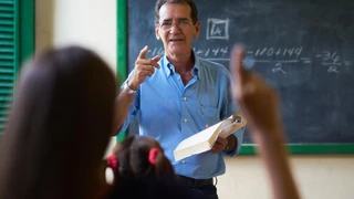 A teacher in a blue shirt engages smilingly with students in a classroom. He's holding a book and pointing, with a chalkboard of math equations behind him.