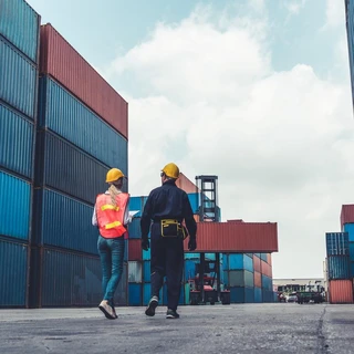 Two workers in safety gear walk through a shipping yard, surrounded by stacked blue and red containers under a partly cloudy sky.