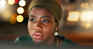 A woman wearing a green headscarf gazes thoughtfully forward. Warm, blurred bokeh lights create a cozy ambiance in the background.