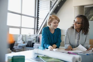 Two women in an office, wearing business attire, are smiling and discussing documents on a table. The atmosphere is collaborative and positive.