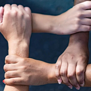 Four interlocked hands with diverse skin tones form a square, symbolizing unity and teamwork, set against a dark, blurred background.