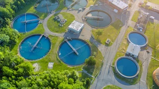 Aerial view of a wastewater treatment plant with eight circular clarifiers filled with water, surrounded by greenery and connected by walkways. Sunny, clear day.