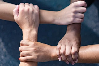 Four interlocked hands, each gripping another’s wrist, form a square against a dark background. The image symbolizes unity and diversity.