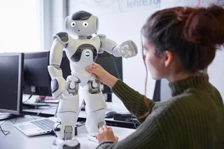 A person interacts with a white humanoid robot marked "NAO" in an office setting. Two computer monitors and a laptop are on the desk, suggesting a tech-focused environment.
