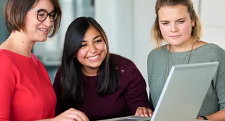 Three women are gathered around a laptop, smiling and engaged in discussion. They appear focused and collaborative, sitting in a bright office space.