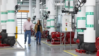 Two people walking through a spacious room featuring prominent red and white pipes along the walls and ceiling.